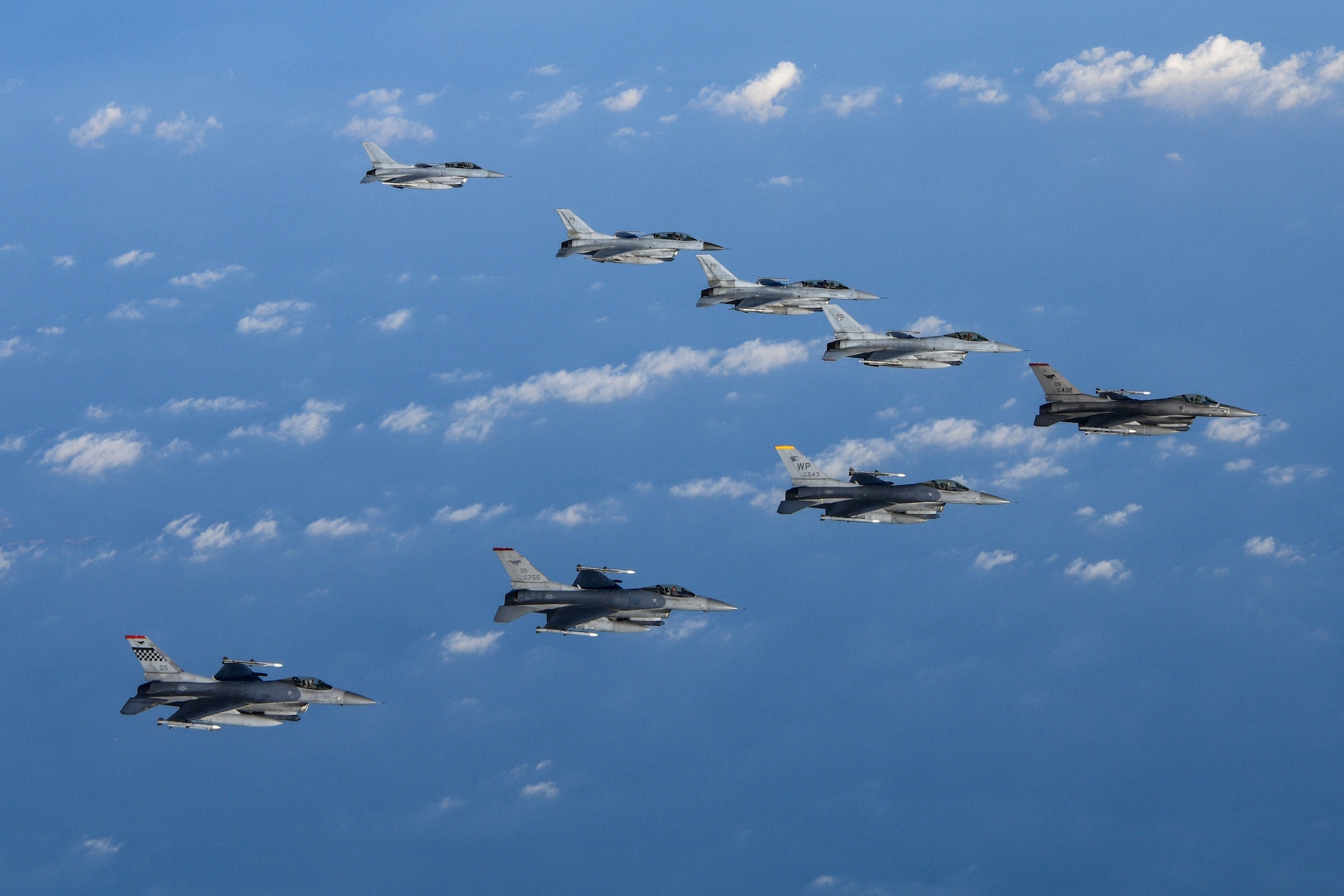Eight F-16 fighter jets fly in a v formation over blue water with white clouds.