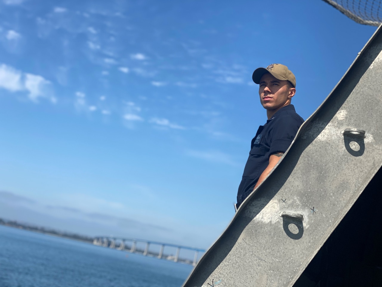 A sailor stands on a military ship at sea with a bridge in the distance.