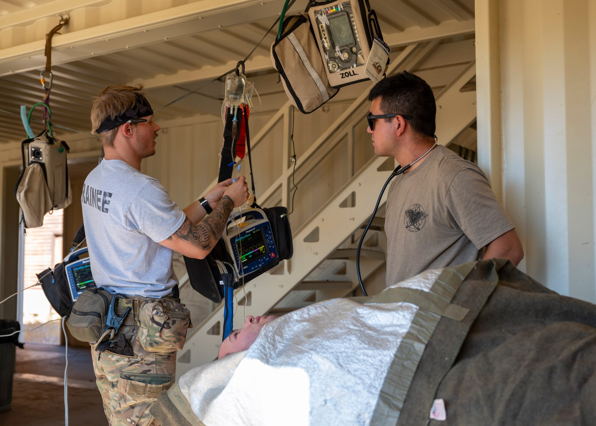Two men in military clothing prepare an IV for a simulated patient who is laying down.