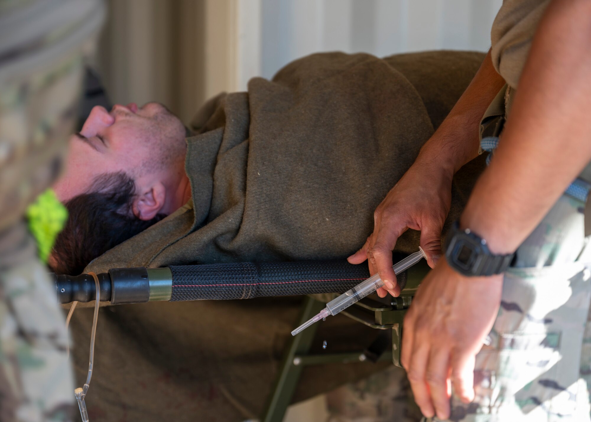 A simulated patient lays down while a man wearing military clothing holds a syringe nearby.