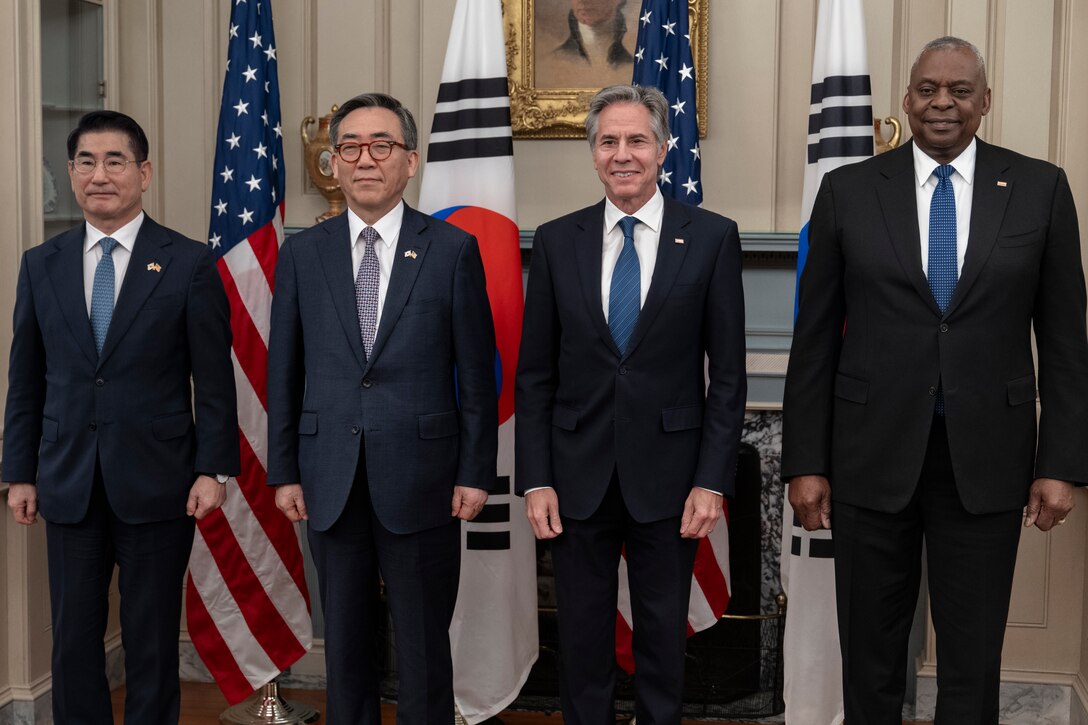 Four government officials stand in front of flags.