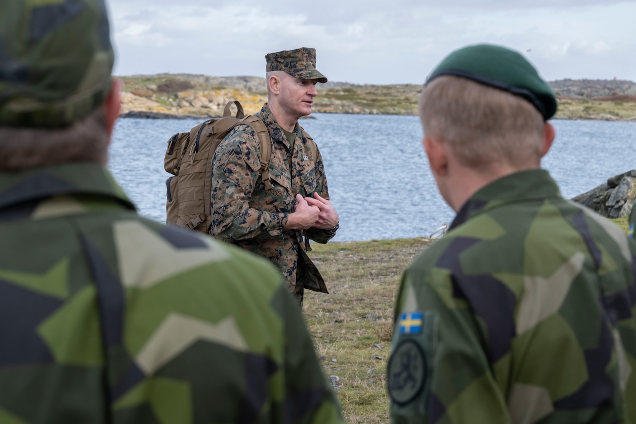 A leader speaks to troops in front of a body of water.