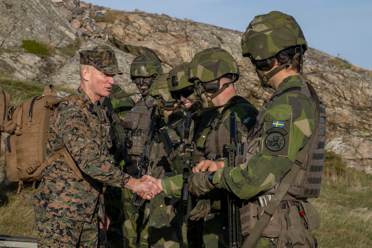 A military leader shakes hands with a group of Marines nearby a rocky mountain.