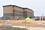 A large building is under construction.  A construction worker, standing behind fencing, moves a piece of wood.