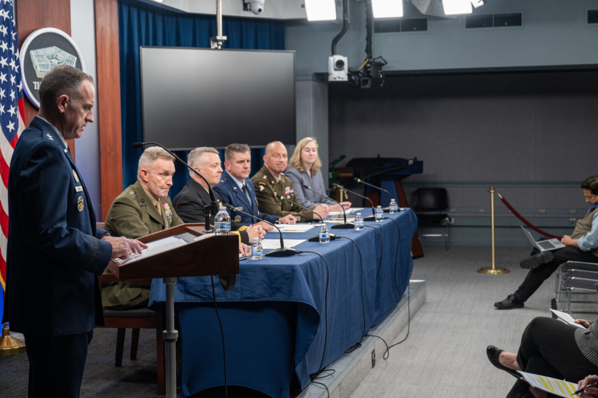 A man in a dress military uniform stands at a lectern with a microphone.  Behind him, four men in dress military uniforms and one woman in civilian clothing are seated at a table with microphones.
