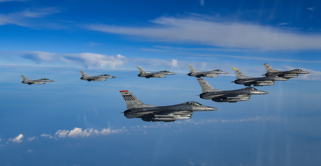 Eight F-16 fighter jets fly in a v formation over blue water with white clouds.