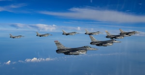 Eight F-16 fighter jets fly in a v formation over blue water with white clouds.