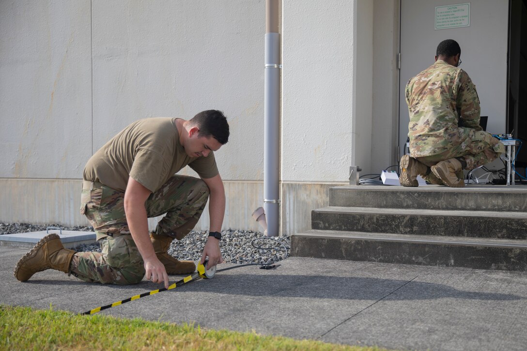 U.S. Air Force Airman 1st Class Syrus Terwilliger, left, a radio frequency transmission systems technician with 374th Communications Squadron, 374th Airlift Wing, 5th Air Force and a native of New Jersey, places caution tape over an exposed wire while Senior Airman Nigel Jessamy, right, a cyber transport technician with 374th Communications Squadron, 374th Airlift Wing, 5th Air Force and a native of Georgia, connects his workstation to the internet during operation Adamantium Shield, at Marine Corps Air Station Iwakuni, Japan, Aug. 21, 2024. Operation Adamantium Shield is designed to simulate establishing communication in a foreign environment to pass information to those in need. (U.S. Marine Corps photo by Lance Cpl. Dahkareo Pritchett)