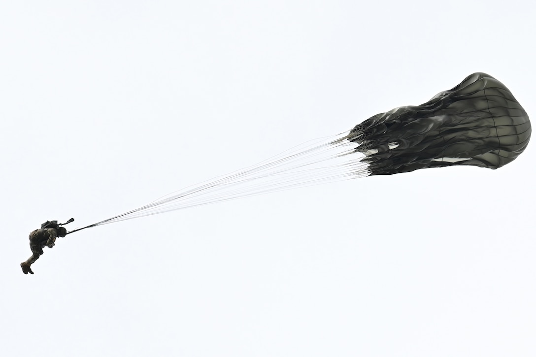 A paratrooper waits for a black parachute to open while descending against a white background.