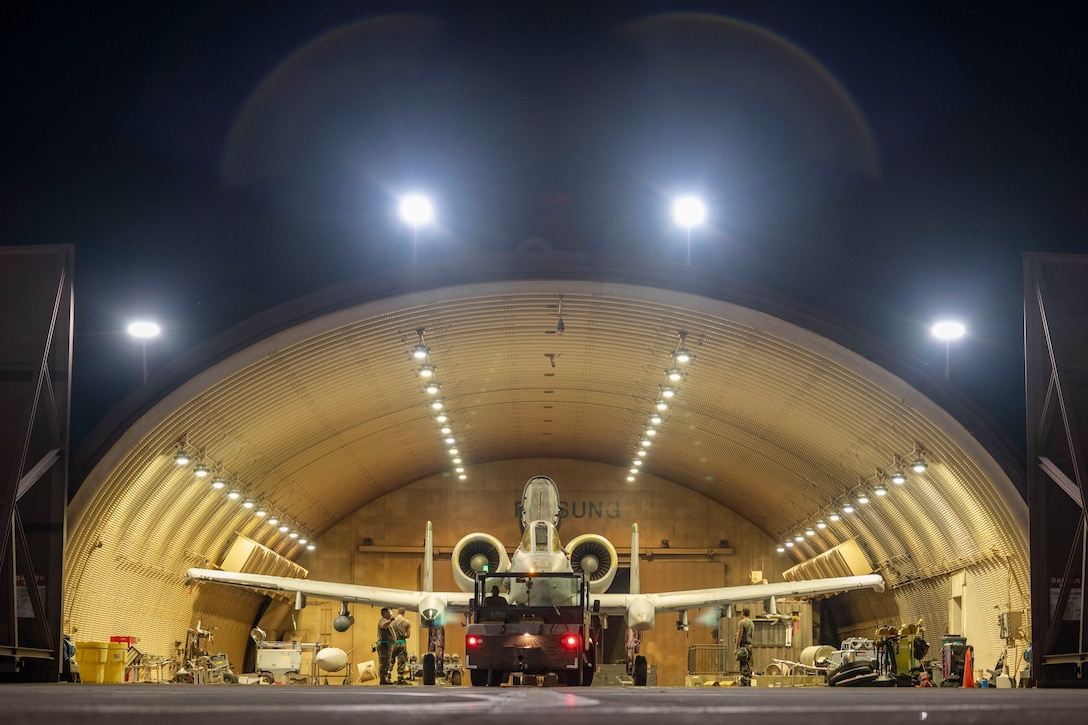 A military aircraft sits in a brightly lit hangar at night as airmen stand around it.