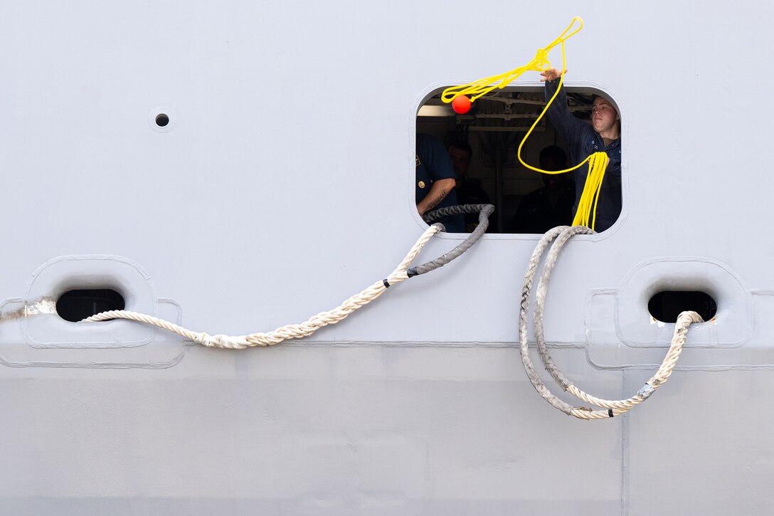 A sailor throws a yellow rope with a weighted red ball from the porthole of a ship. Other thick white ropes are seen against the side of the ship.