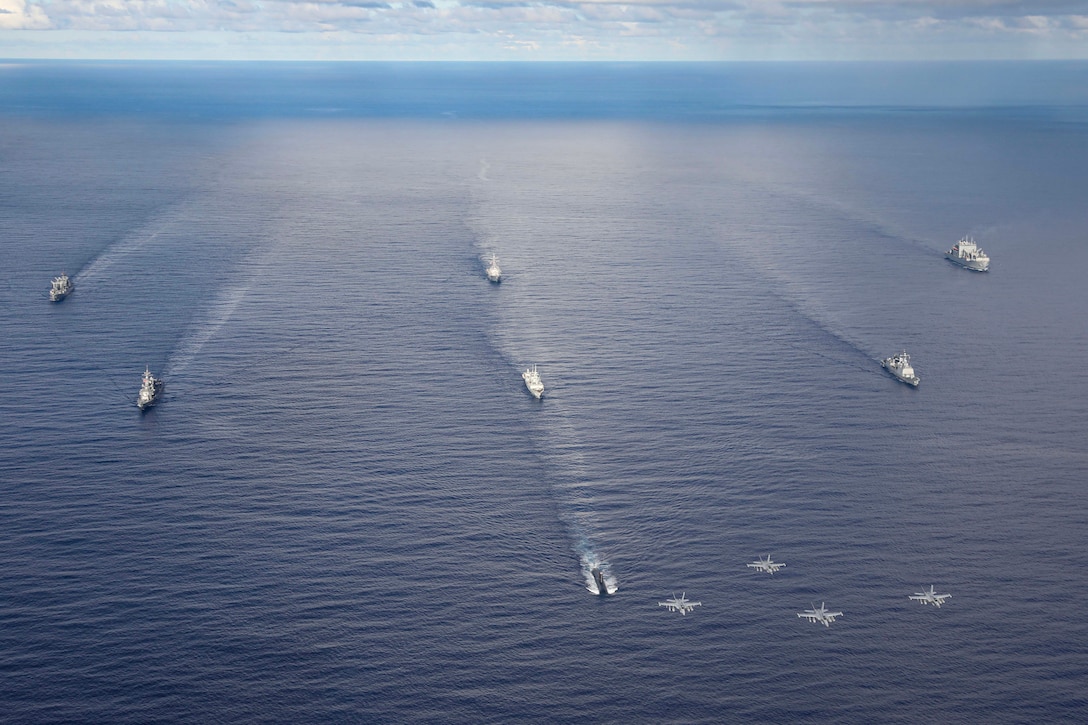 Seven military ships sail in open water as four military aircraft fly above during daylight.