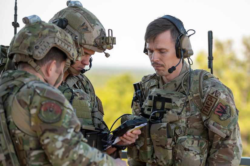 Service members wearing battle gear stand together and look at a hand-held device.