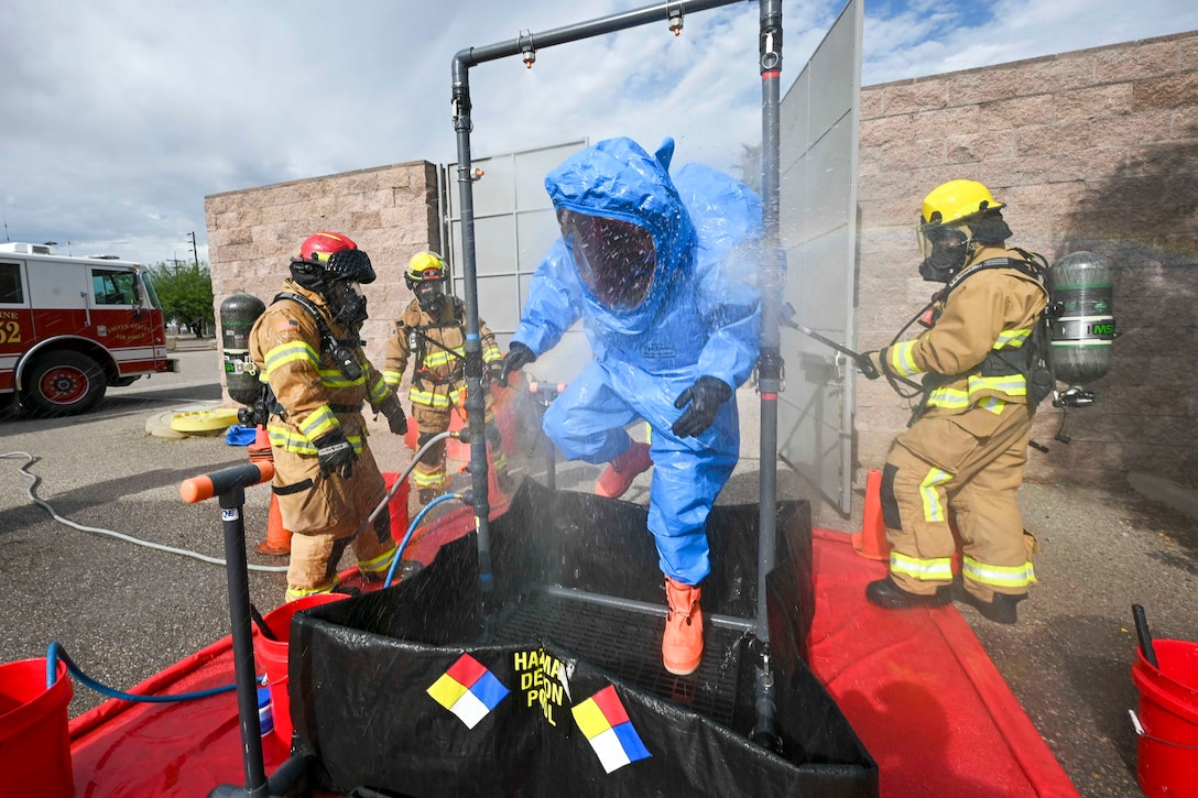 An airman wearing a blue hazmat suit stands in a makeshift shower as airmen in firefighting suits spay them with water.