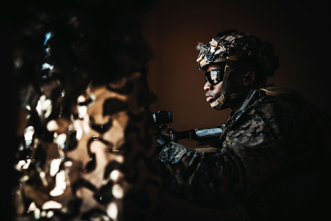 A Marine aims a weapon while looking through mesh netting in a dark area.