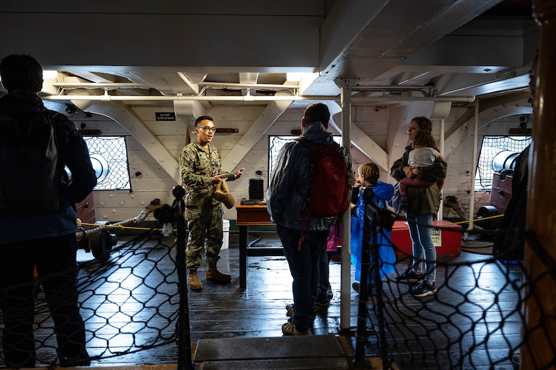 A sailor speaks to a group of people and children below deck on an old ship.