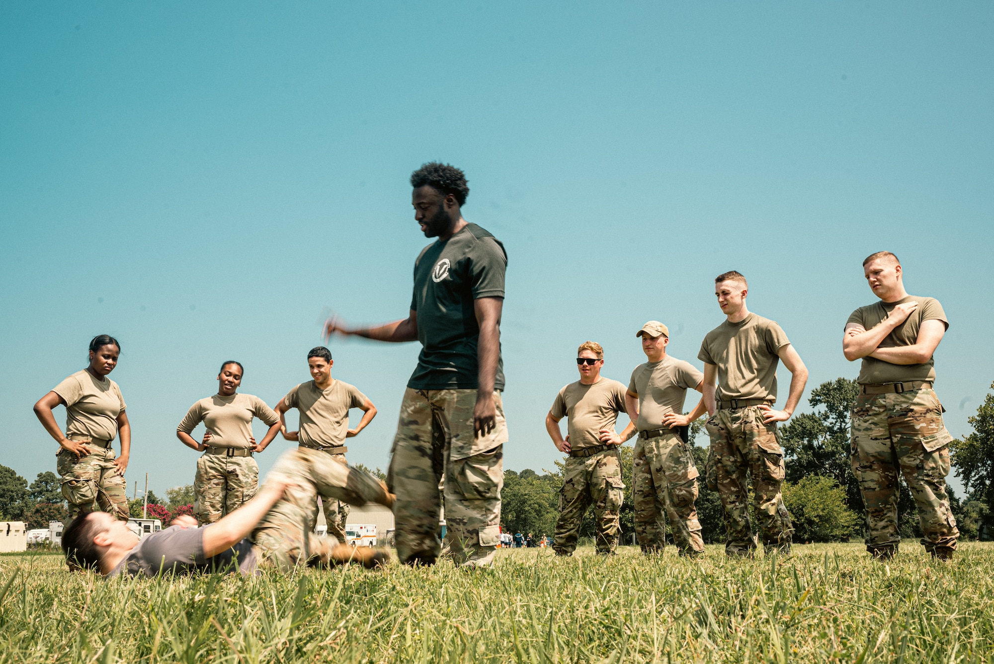 A combat instructor in a black shirt teaches airmen combatives that are standing around him