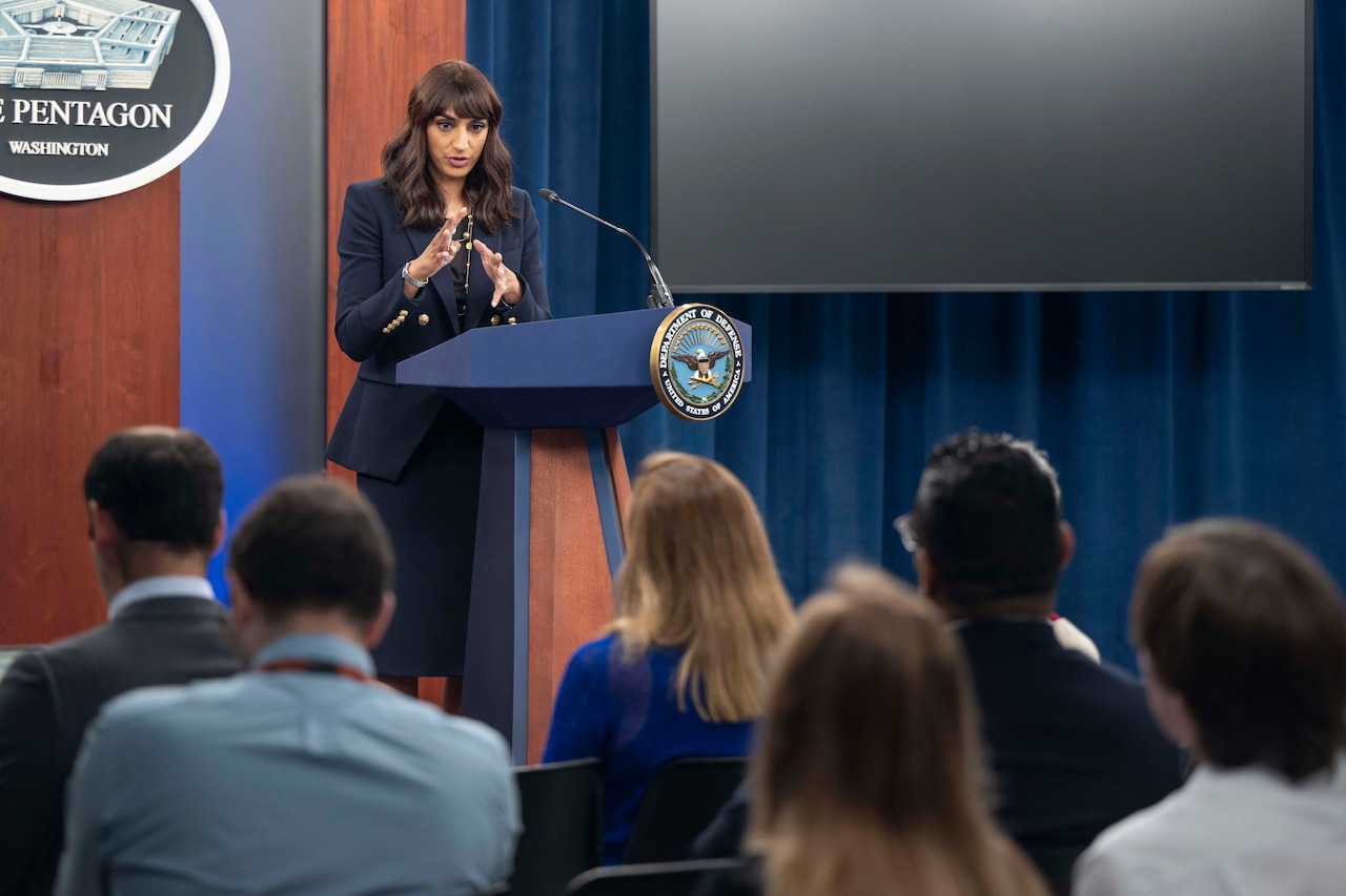 A spokesperson speaks to an audience from a lectern with the Pentagon seal and a large blank montor in the background.