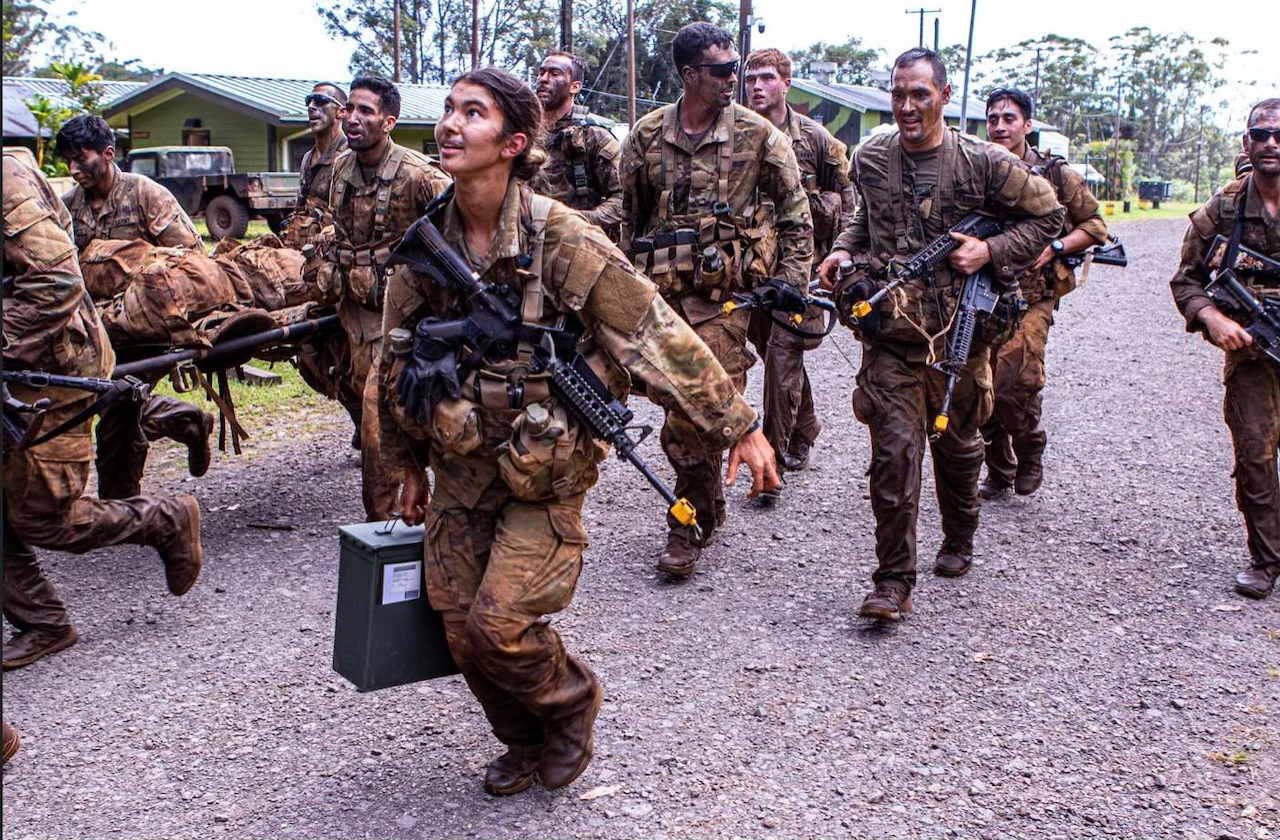 Soldiers run with equipment outside on a gravel path during the day.