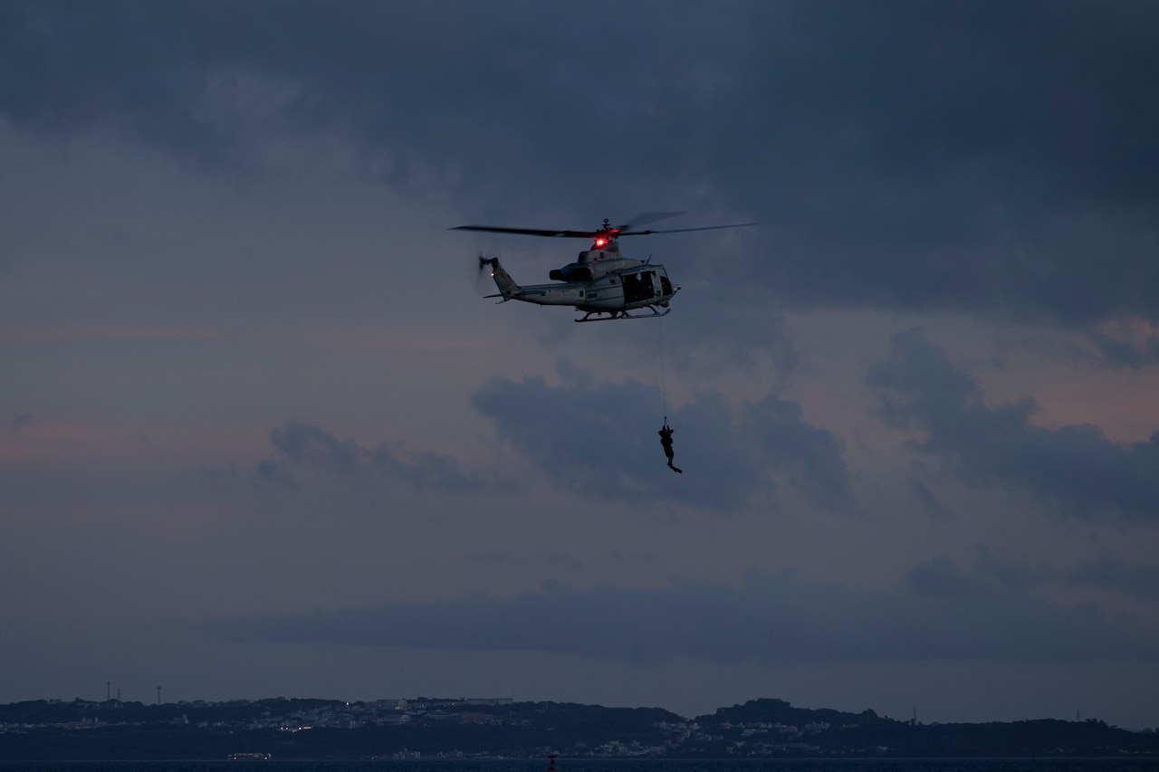 A Marine hoists onto a UH-1Y Venom helicopter in the dark over a body of water.