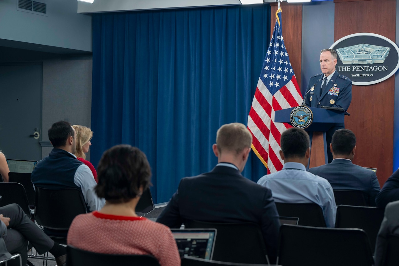 A military officer stands behind a podium and speaks to people sitting in chairs.