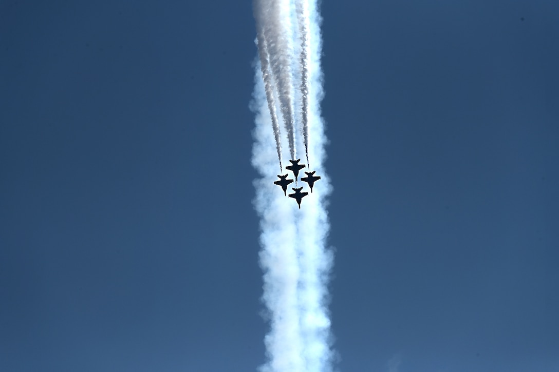 Four military aircraft are photographed from below flying in formation, leaving a vapor trail against a bright blue sky.