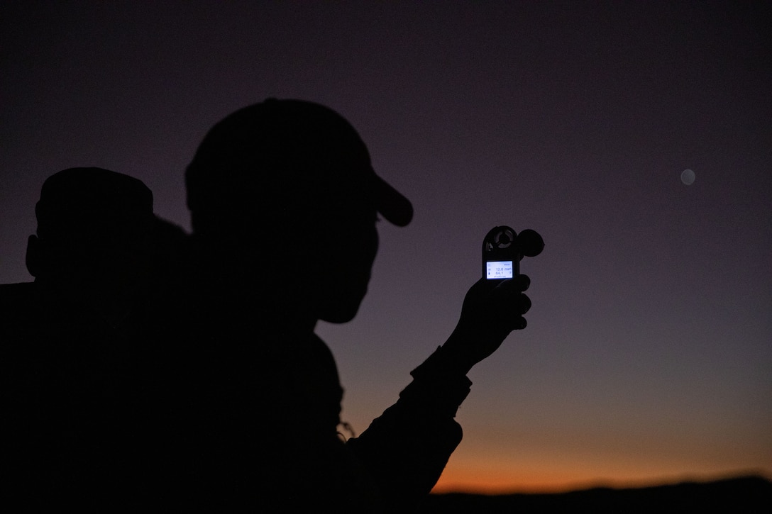 A soldier wearing a cap is seen in silhouette checking the digital screen on a handheld wind speed device at twilight.