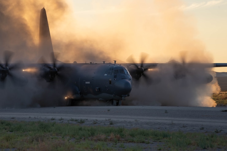 A military aircraft taxis on a dirt runway, blowing up dust.