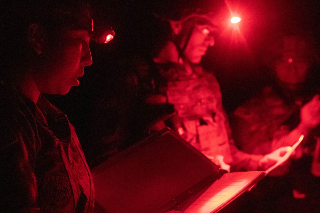 Three soldiers wearing headlamps read through binders in the dark illuminated by red light.