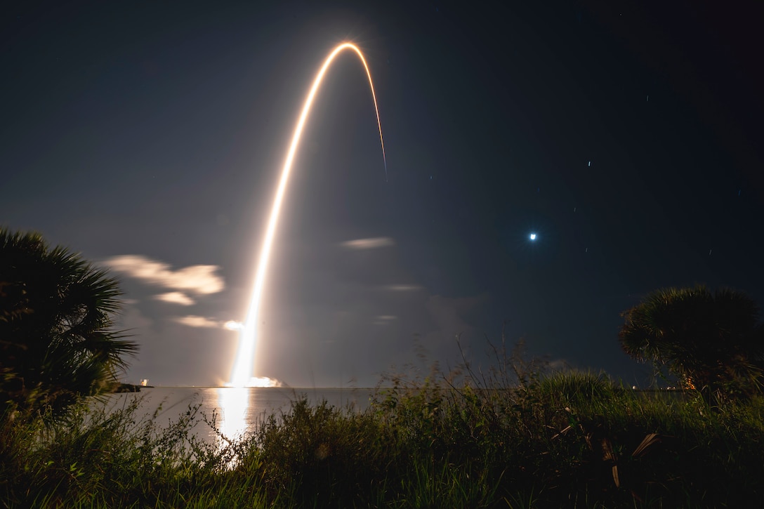 A rocket launches in the dark leaving a streak in the sky with grass, trees in the foreground.