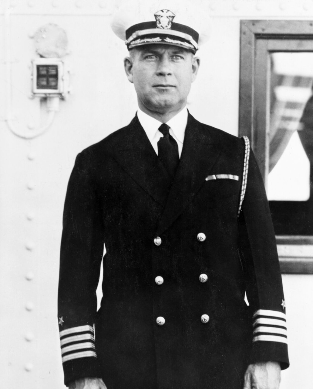 A Navy officer stands on a ship wearing an officer uniform and cap.