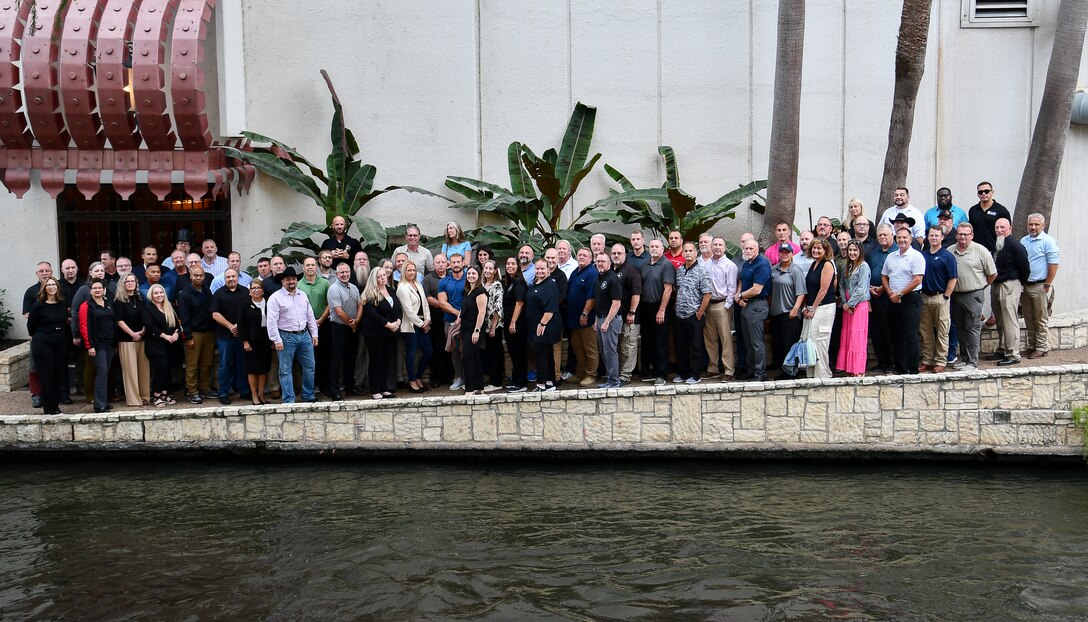 A large groups of people stand in front of water with a wall behind them. there are also a few plants behind them.