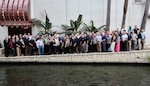 A large groups of people stand in front of water with a wall behind them. there are also a few plants behind them.