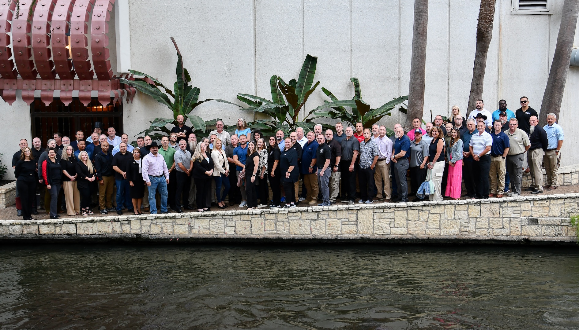 A large groups of people stand in front of water with a wall behind them. there are also a few plants behind them.