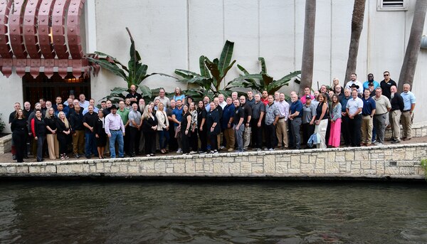 A large groups of people stand in front of water with a wall behind them. there are also a few plants behind them.
