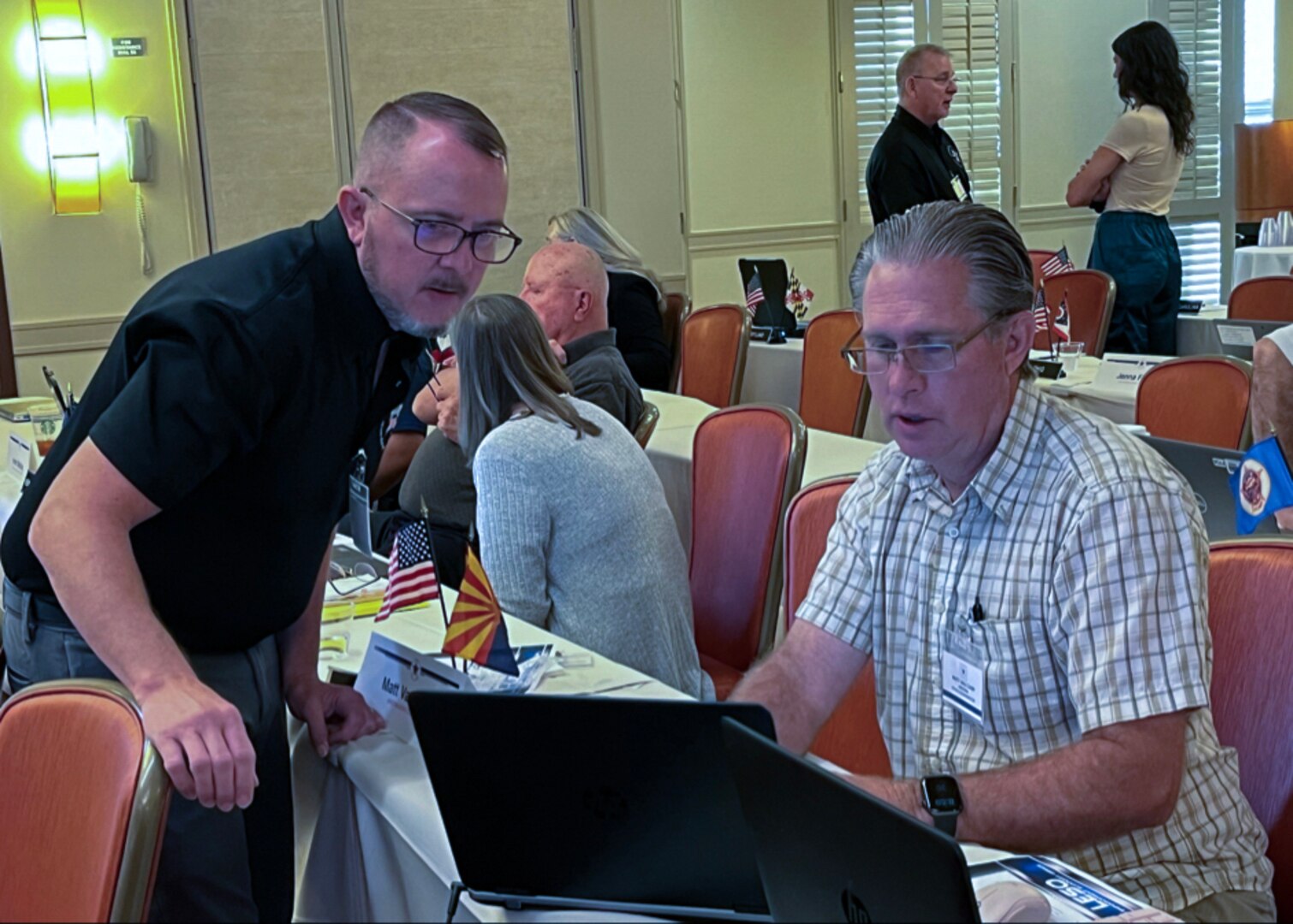 A man in a blue shirt stands and looks at a laptop monitor while a seated man in a striped shirt does the same.