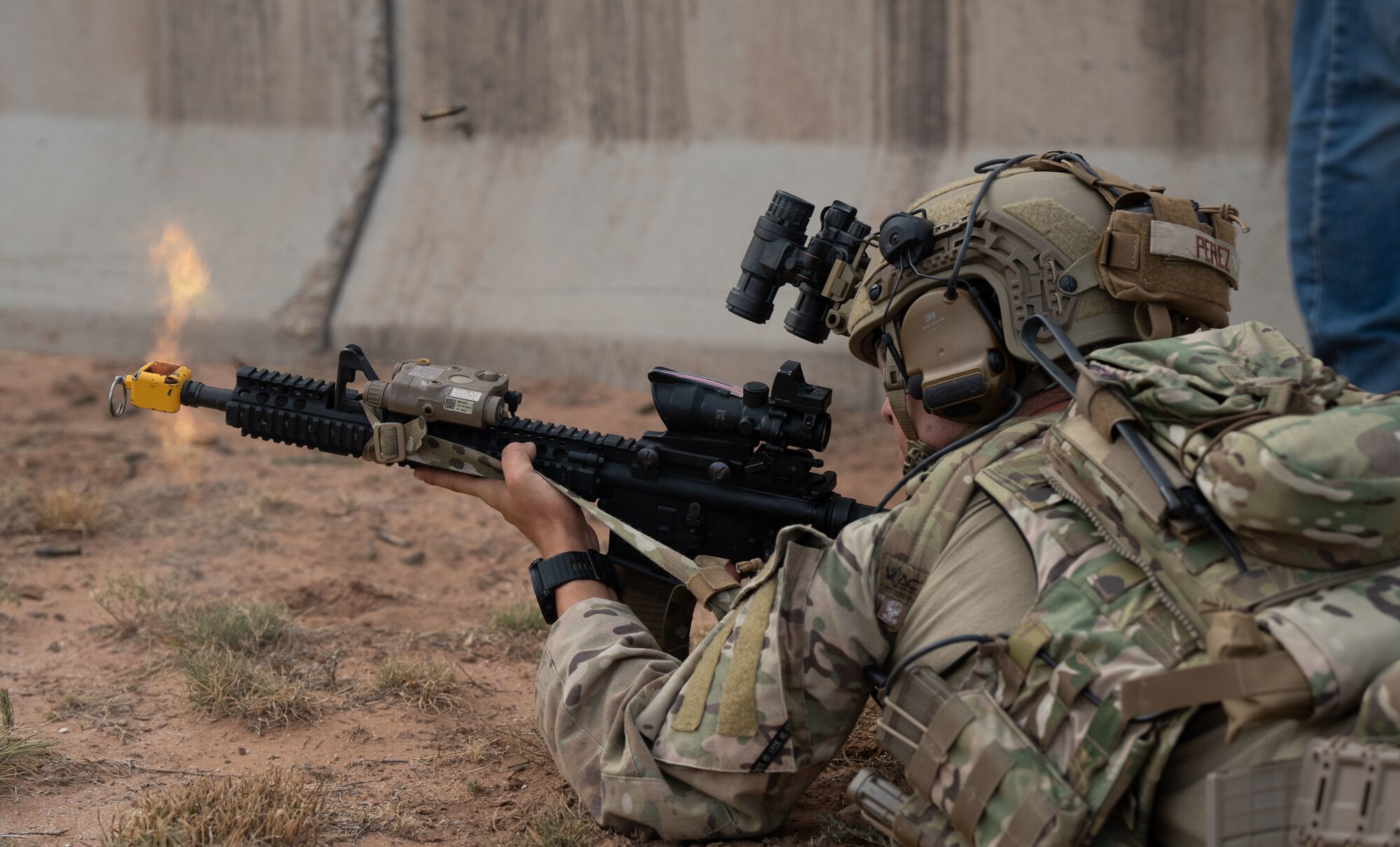 A man in military uniform and military equipment lays in the prone firing a rifle.