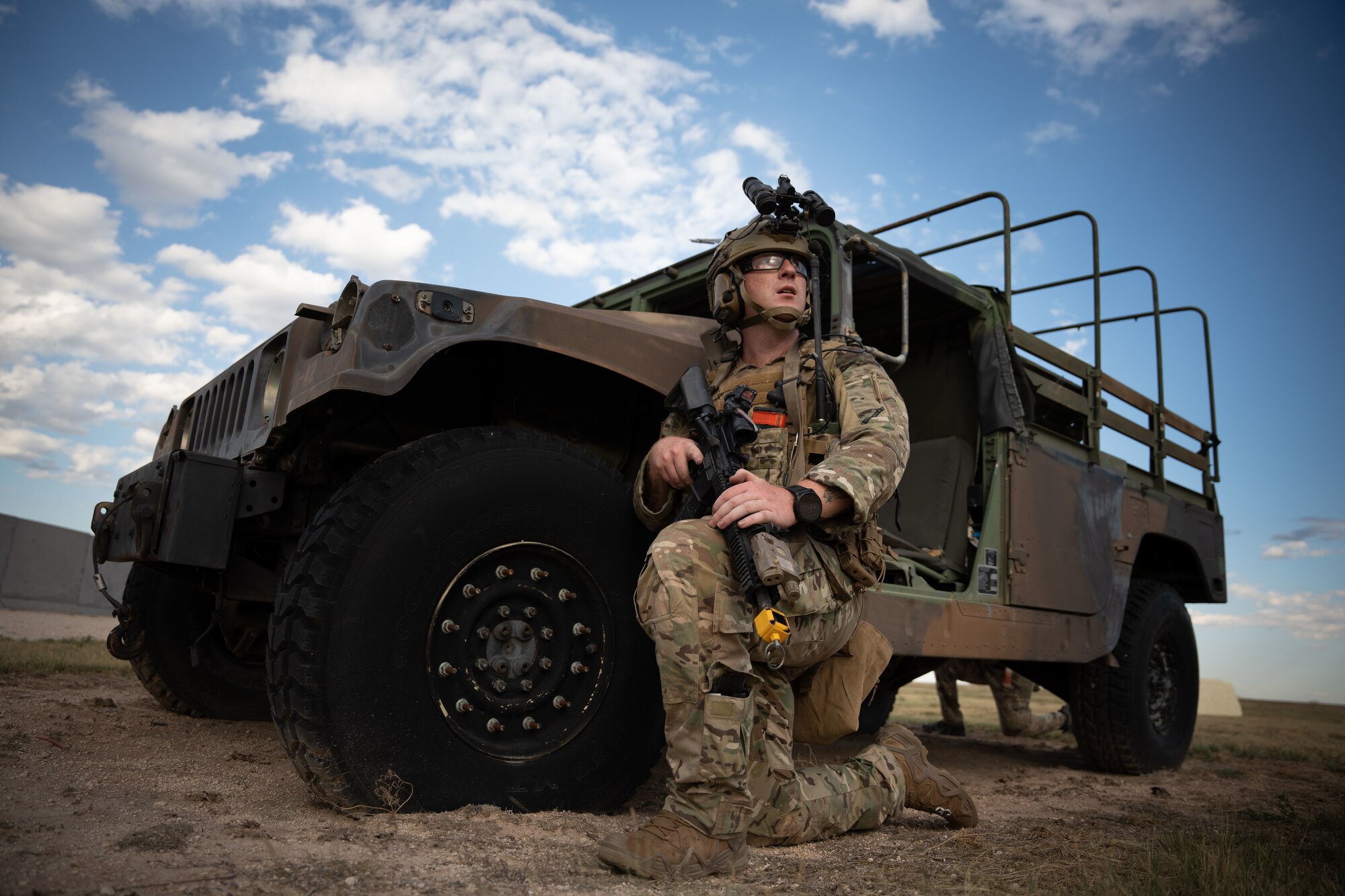 A man in military uniform, military equipment, and armed with a rifle kneels next to a military vehicle and looks in the distance.