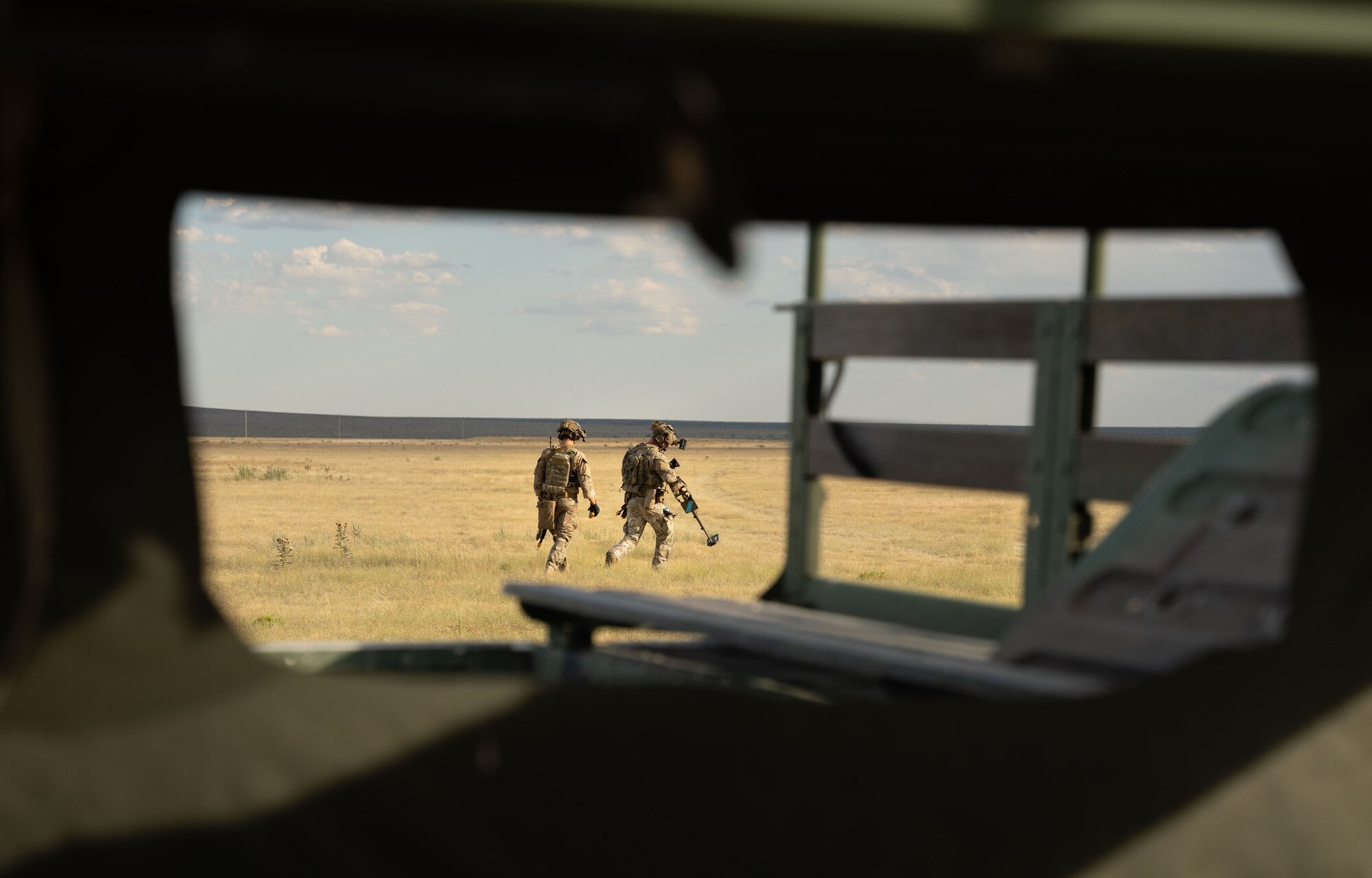 Two men in military uniform swing military equipment along the ground in a field.