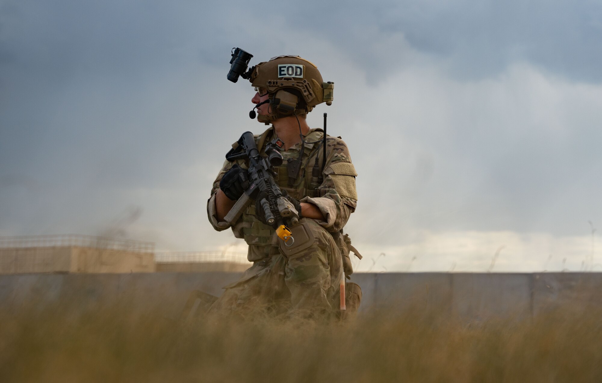 A man in military uniform, military equipment, and armed with a rifle knees in a field looking into the distance.