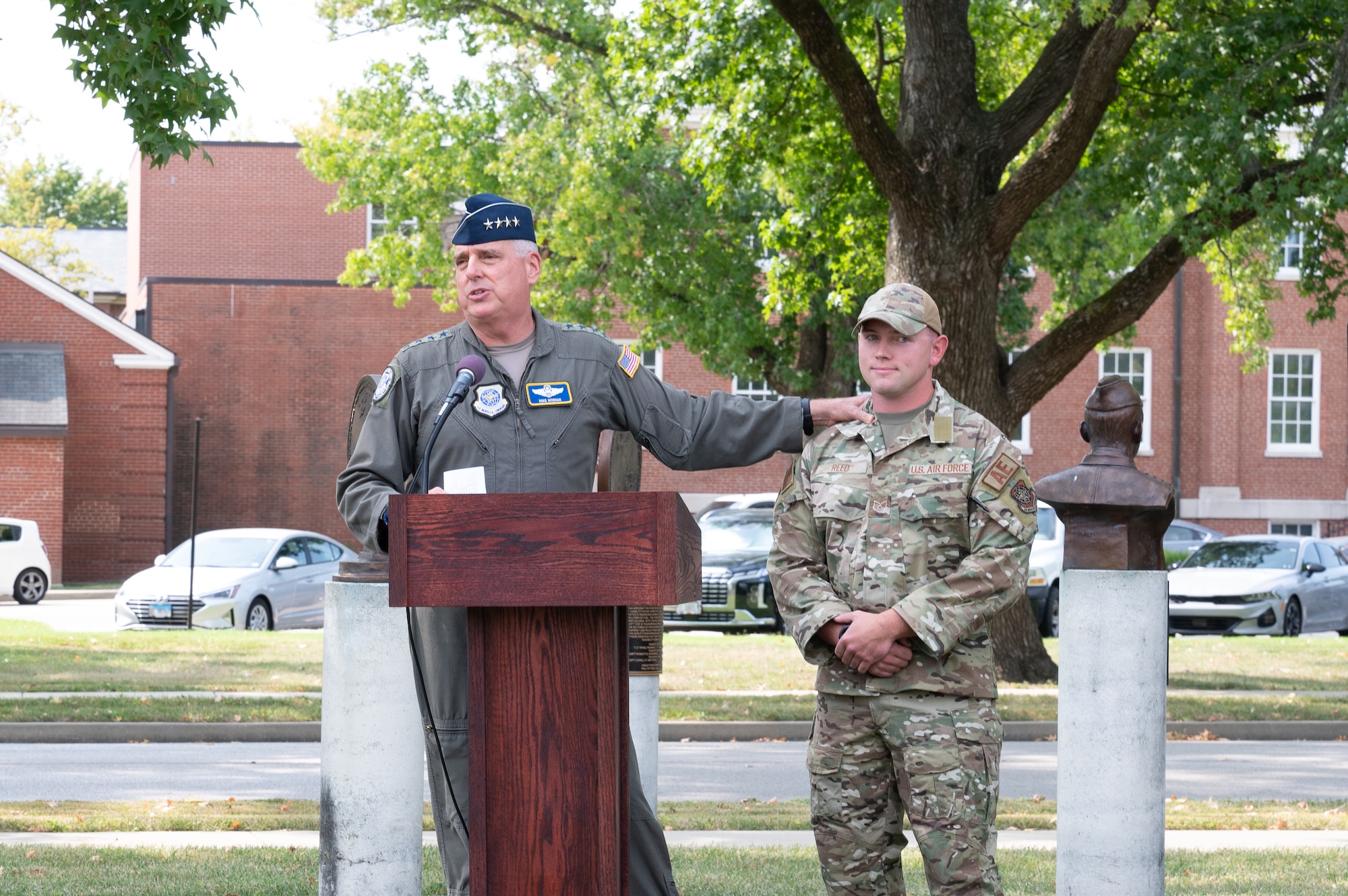 U.S. Air Force Gen. Mike Minihan stands at a podium with his hand resting on Staff Sgt. Morgan Reed's shoulder.