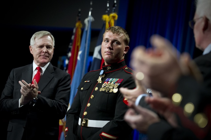 A service member stands on a stage while two others nearby applaud.