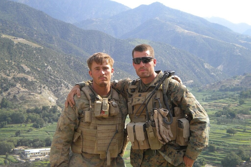 Two service members in combat gear pose for a photo on a mountainside.