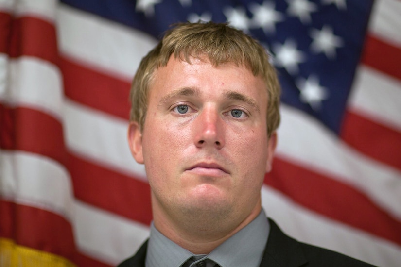 A person looks directly into the camera for a portrait with the American flag in the background.