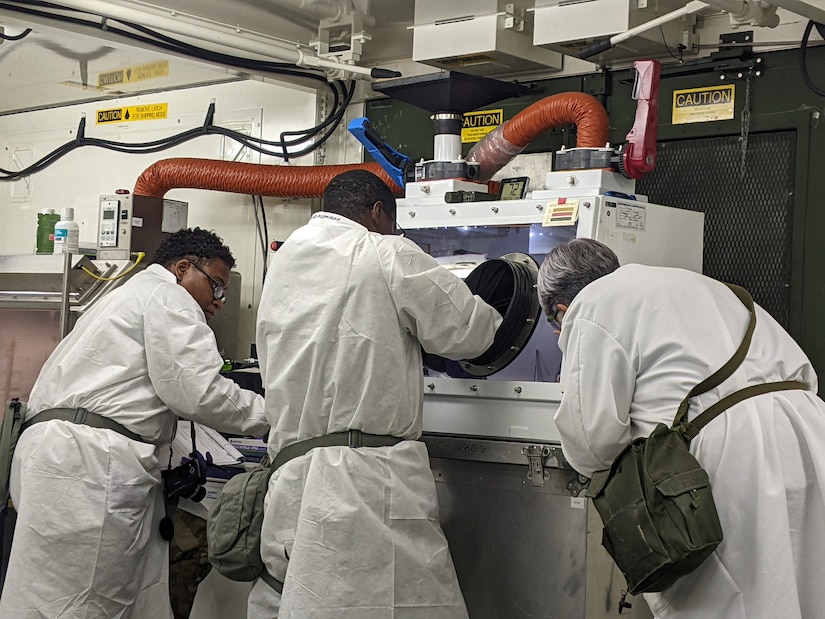 Three scientists in white coats lean in over equipment inside a laboratory.