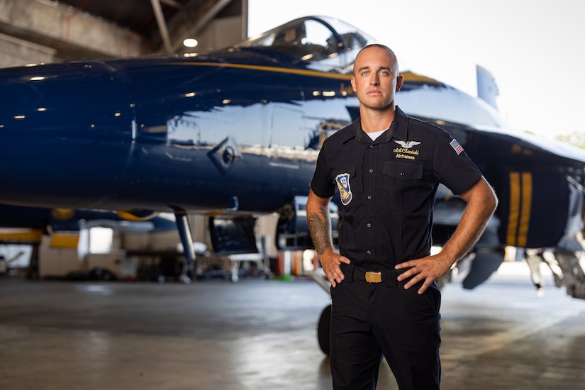 A pilot poses in front of a blue jet aircraft inside a hangar.