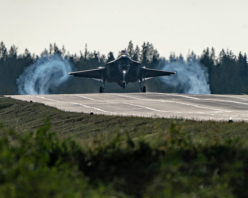 A fighter jet lands on a landing strip during the day in front of a forest.