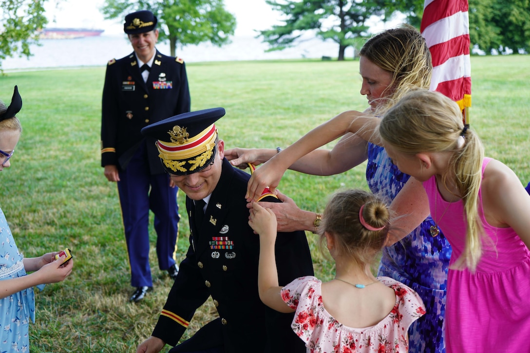 A military officer kneels while family members place new insignia on their formal uniform. Another service member watches in the background.