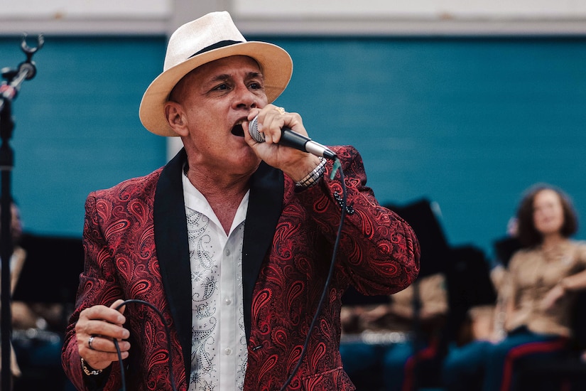 A singer in a red suit and white hat sings inside a building with the Marine Corps band behind him.