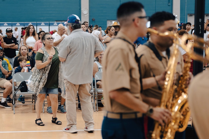 A group of Marines plays instruments while people dance in the background inside a gymnasium.